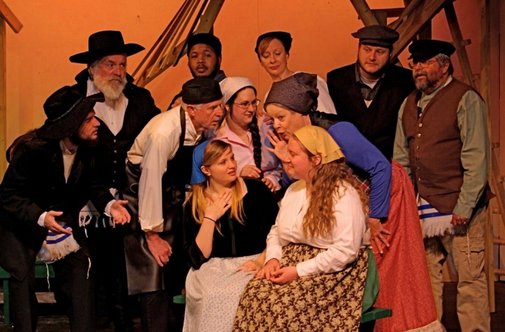  The villagers (L to R seated: Alyson Farnell and Kylie Sommer, standing second row: Jusden Messick, Bob Chauncey, Kelly Ostazeski, and Kathy Jones, third row: Michael Whitehill, Stefan Tisdale, Heather Joyce-Byers, Will Covington and Jim Johnson) are astounded by rapidly-spreading rumors.  